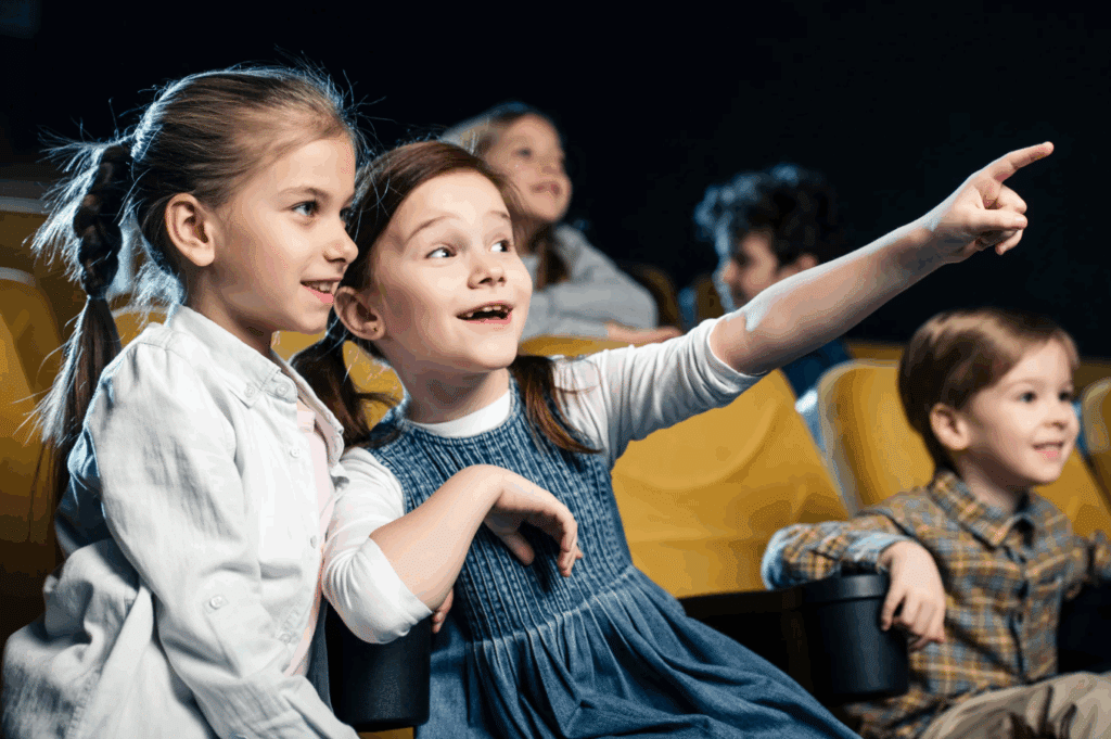 excited kid pointing with finger while watching movie in cinema together with multicultural friends